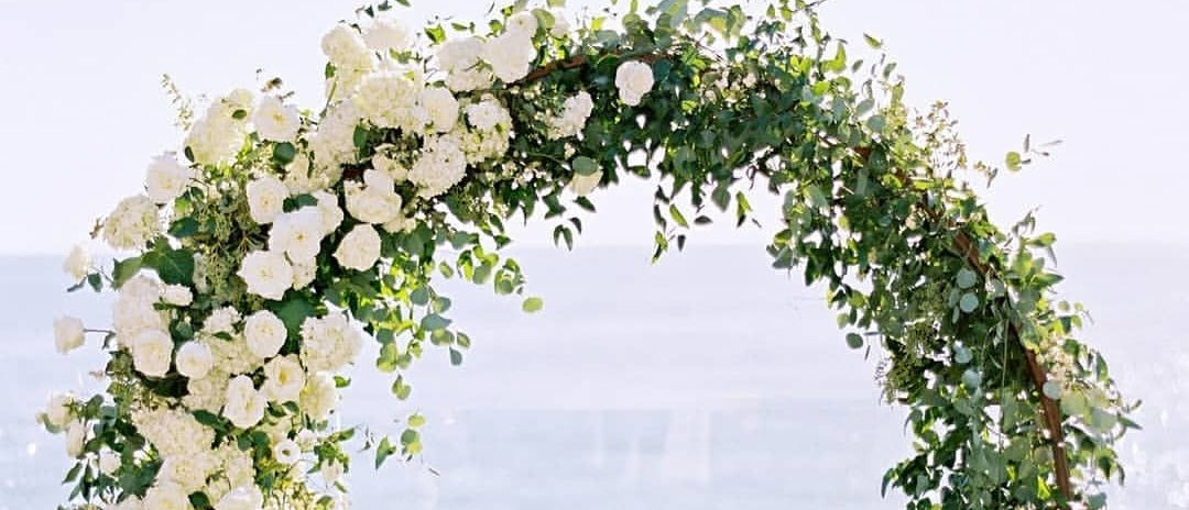 Round white wedding arch with white flowers on green grass in front of ocean 