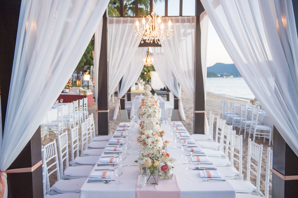 Beach wedding setup with White chiffon drapes, long whit table with white tiffany chairs, white napkins, blush table runners