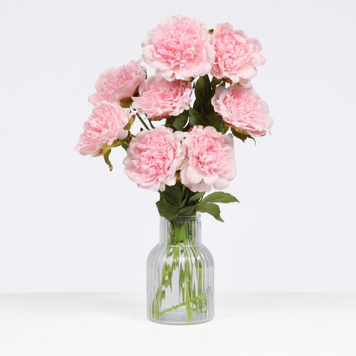 A bouquet of artificial light pink peony flowers with green stems and leaves, arranged in a clear ribbed glass vase against a plain white background.