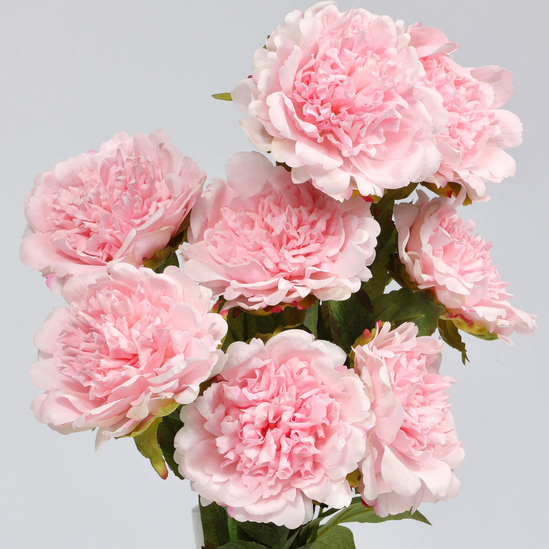 A bouquet of artificial light pink peony flowers with green leaves, arranged against a solid light gray background.