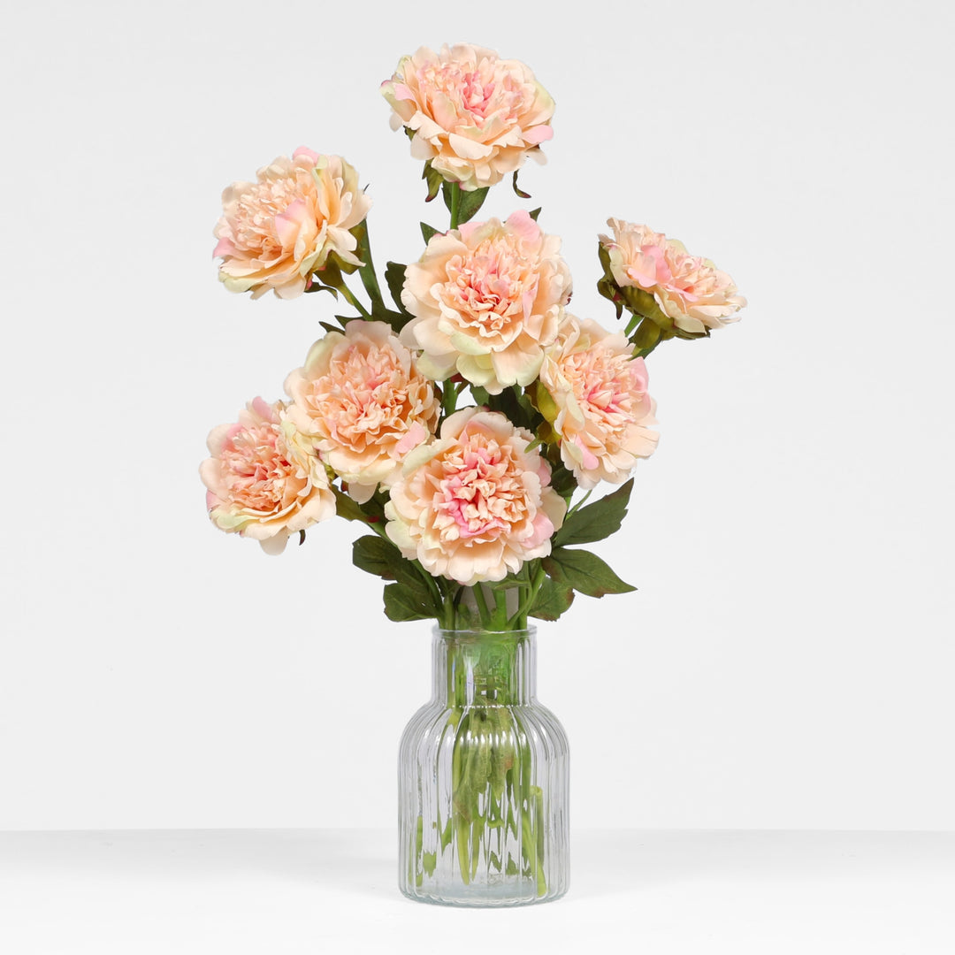 A bouquet of artificial peach peony flowers with green stems, arranged in a clear ribbed glass vase against a light gray studio background.