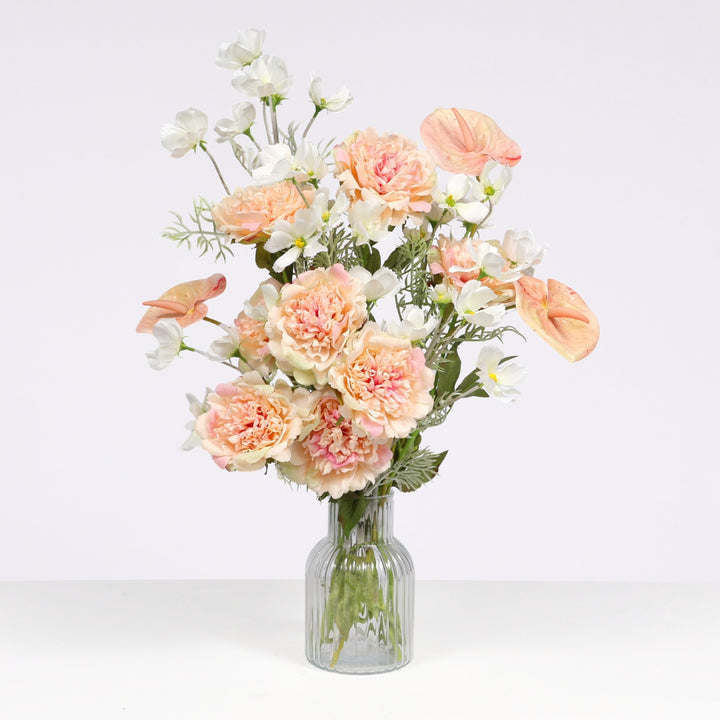 A bouquet of artificial flowers in soft peach, blush pink, and white tones with green foliage, arranged in a clear ribbed glass vase against a light gray studio background.