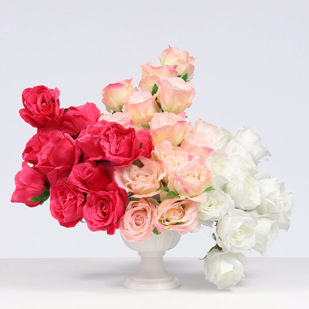 A decorative arrangement of artificial roses in three color groups: vibrant hot pink, soft peach-pink, and white, presented in a small white pedestal vase against a white studio background.
