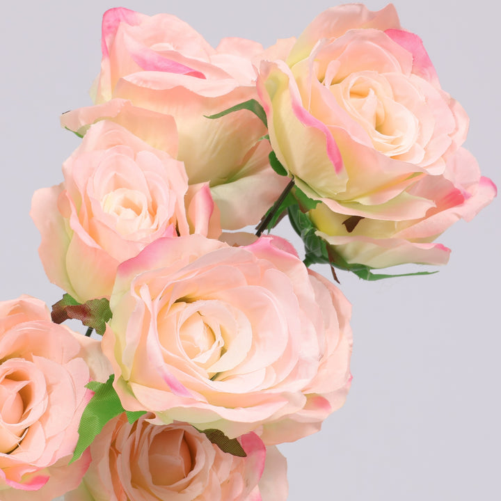 A close-up of several artificial roses in soft blush pink and cream tones with green leaves, arranged against a light gray background.