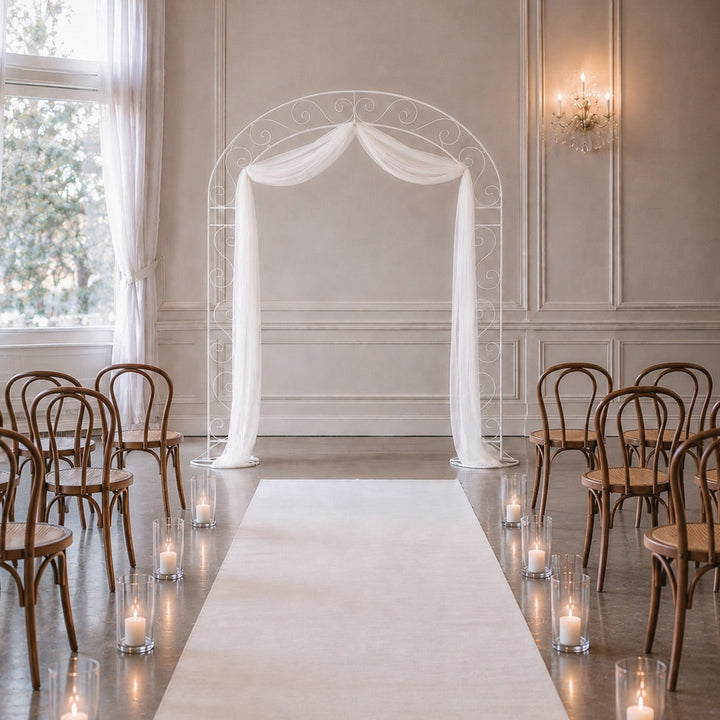 A wedding venue setup with a white vintage frame, draped with white chiffon fabric, a long white aisle runner and wooden chairs arranged on both sides. Small lit candles in glass holders line the aisle.