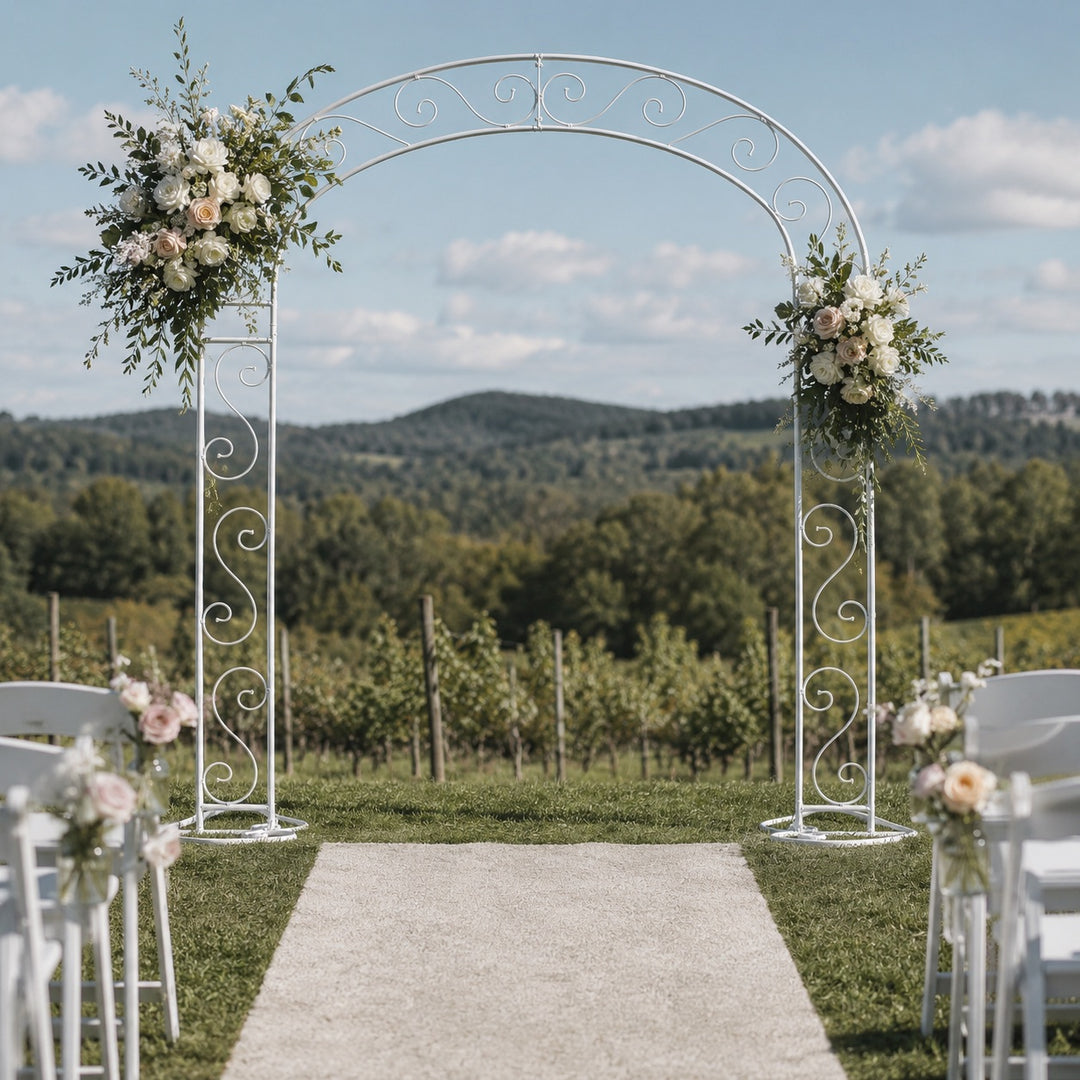 A white vintage scroll wedding arch with floral arrangements on either side, set outdoors on a grassy lawn with an aisle leading through white chairs toward a vineyard under a blue sky.