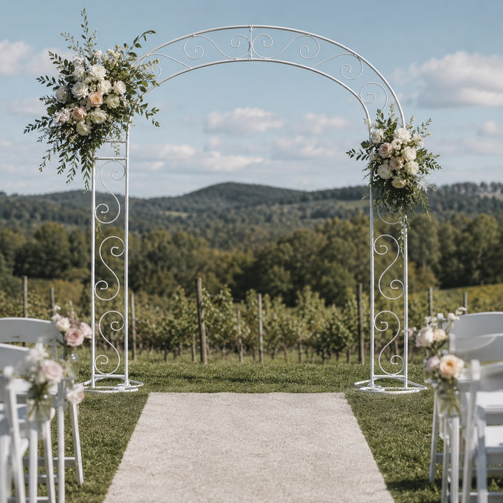 A white vintage scroll wedding arch with floral arrangements on either side, set outdoors on a grassy lawn with an aisle leading through white chairs toward a vineyard under a blue sky.