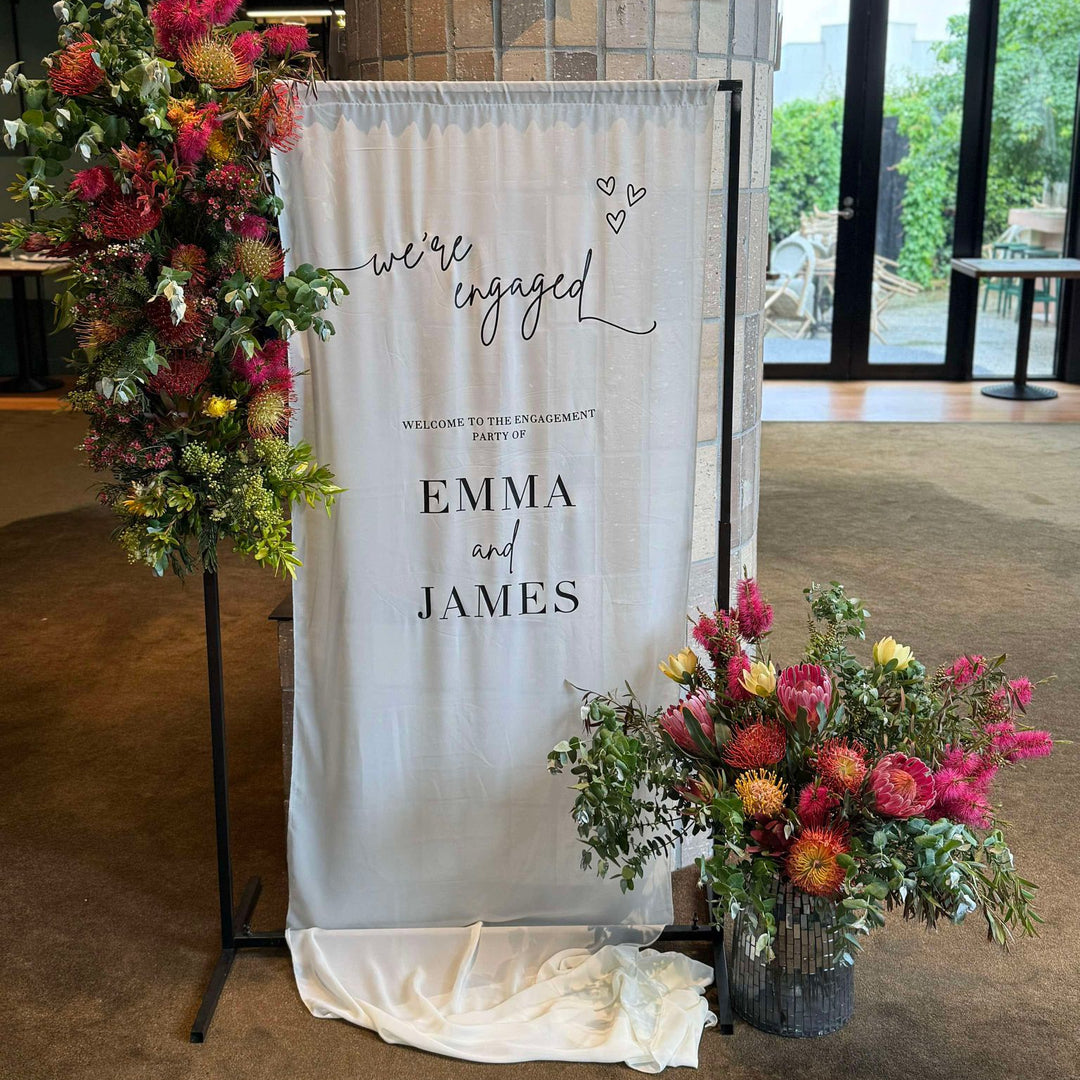 Black height adjustable frame with chiffon engagement sign and native Australian flowers around it. 