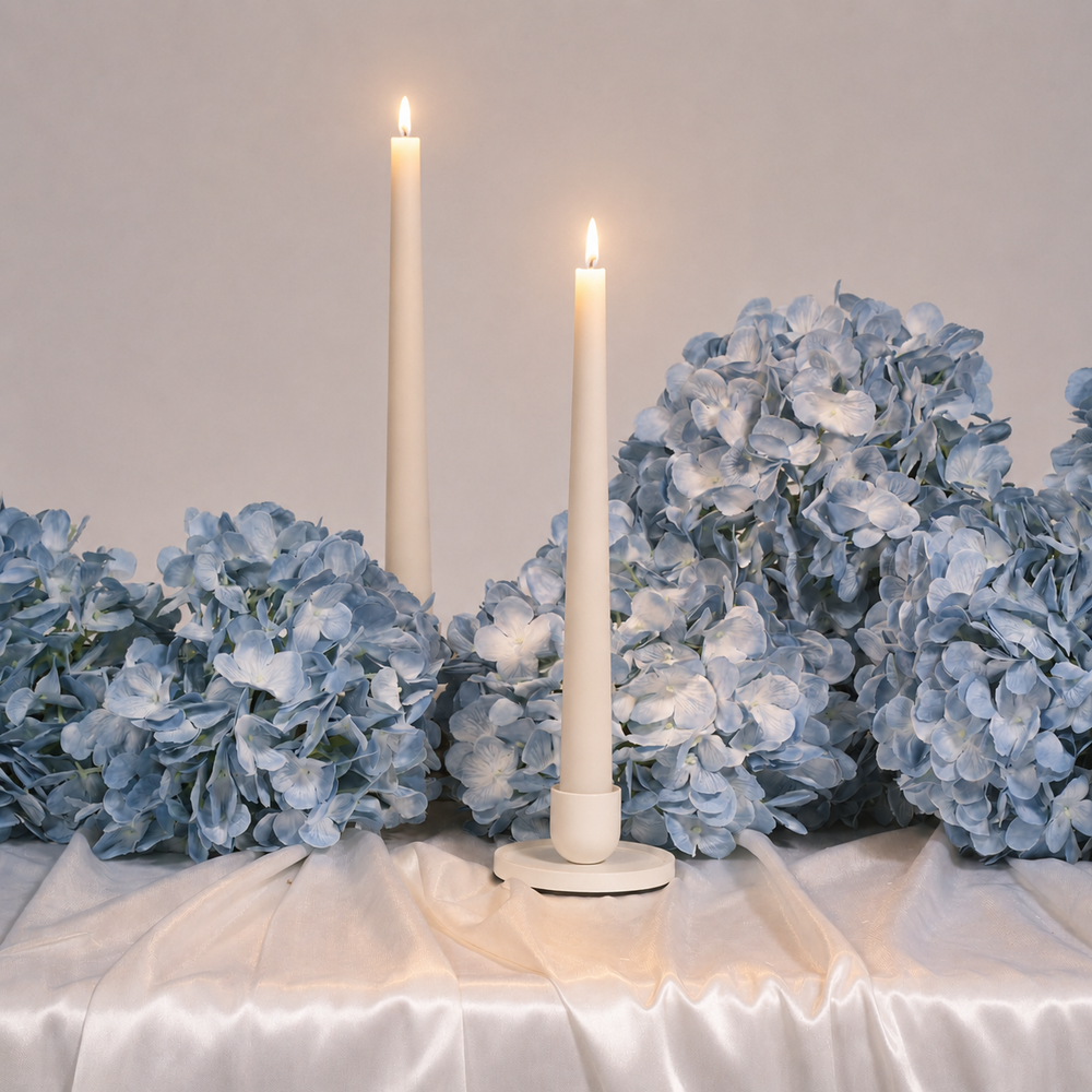 Two white candles in holders surrounded by blue hydrangeas on a white surface.