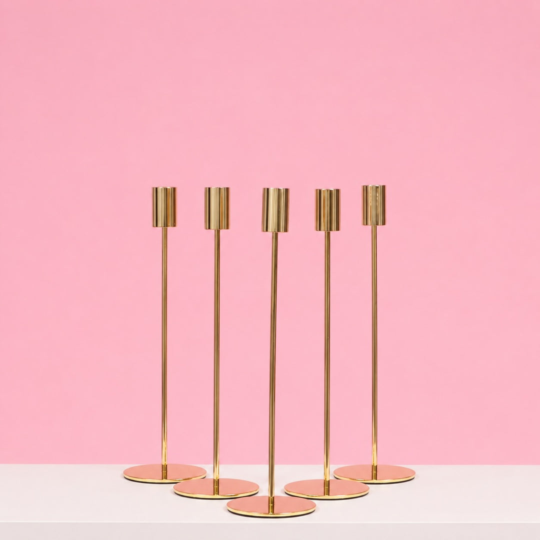 Set of five gold candle holders on a white surface with a pink background