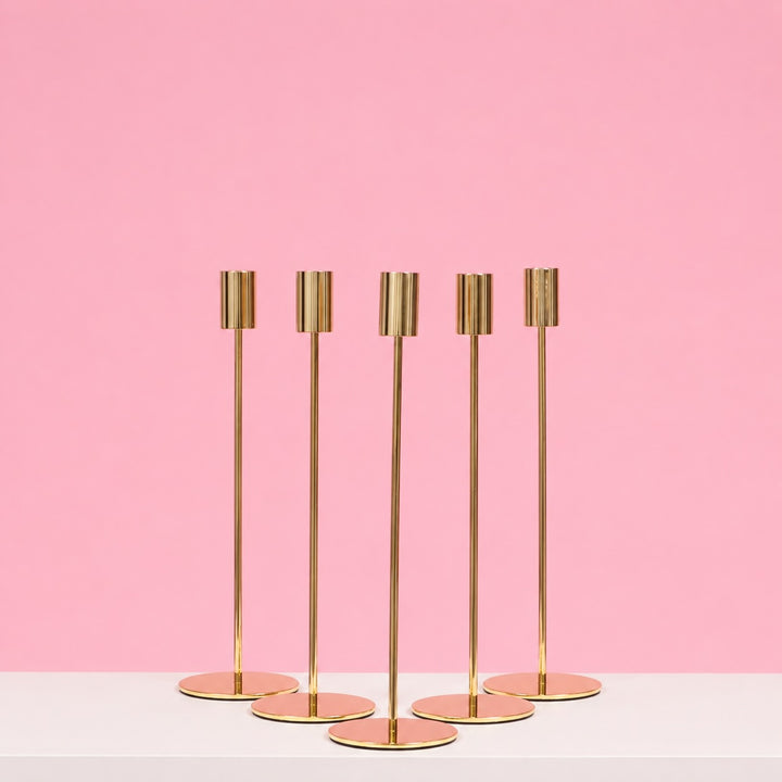 Set of five gold candle holders on a white surface with a pink background