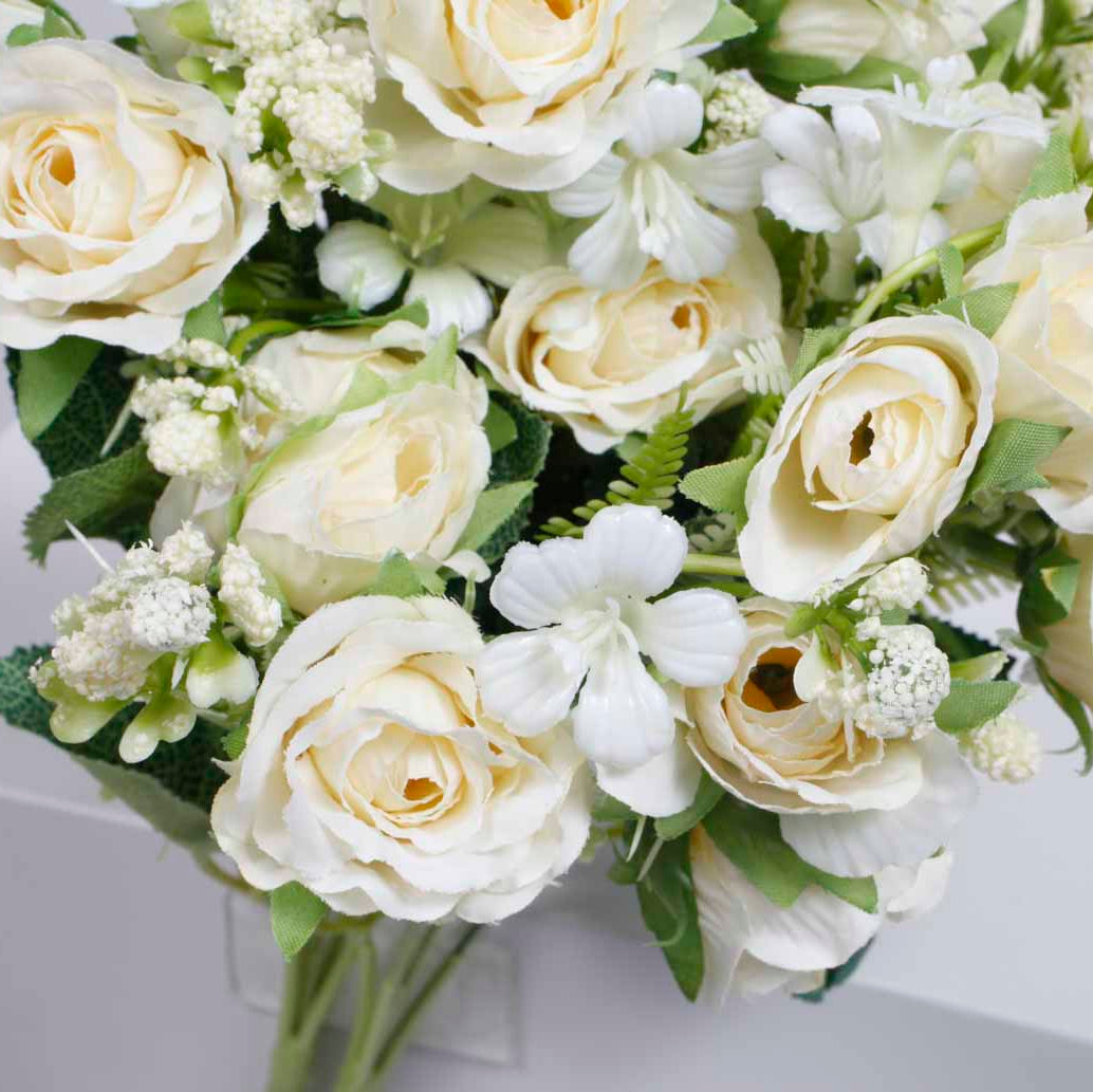 A bouquet of artificial white and cream roses with green leaves and small white filler flowers, arranged in a white box against a light gray background.