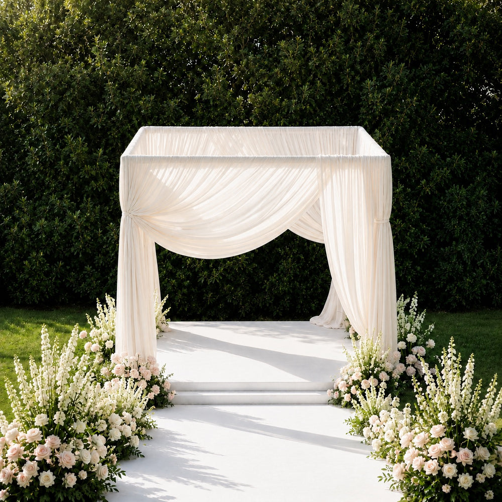 An elegant outdoor wedding altar setup featuring a square canopy draped in flowing ivory fabric, with a white aisle runner leading up steps to the center. The canopy is framed by lush green hedges and decorated with abundant white and blush pink floral arrangements on both sides.