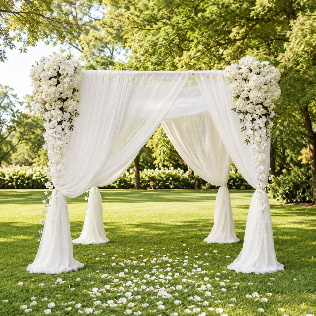 A white draped wedding canopy decorated with large white floral arrangements on the corners, set on a green lawn with scattered white petals and trees in the background.