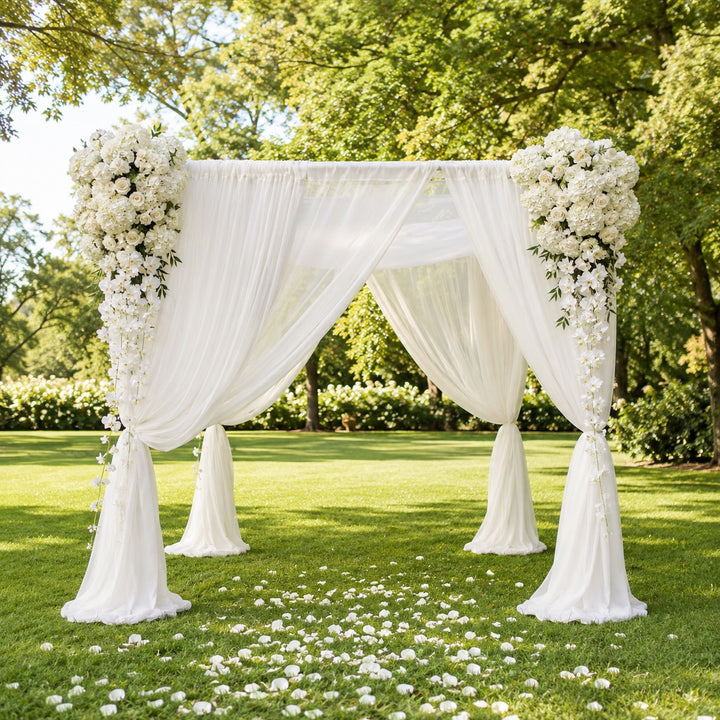 A white draped wedding canopy decorated with large white floral arrangements on the corners, set on a green lawn with scattered white petals and trees in the background.