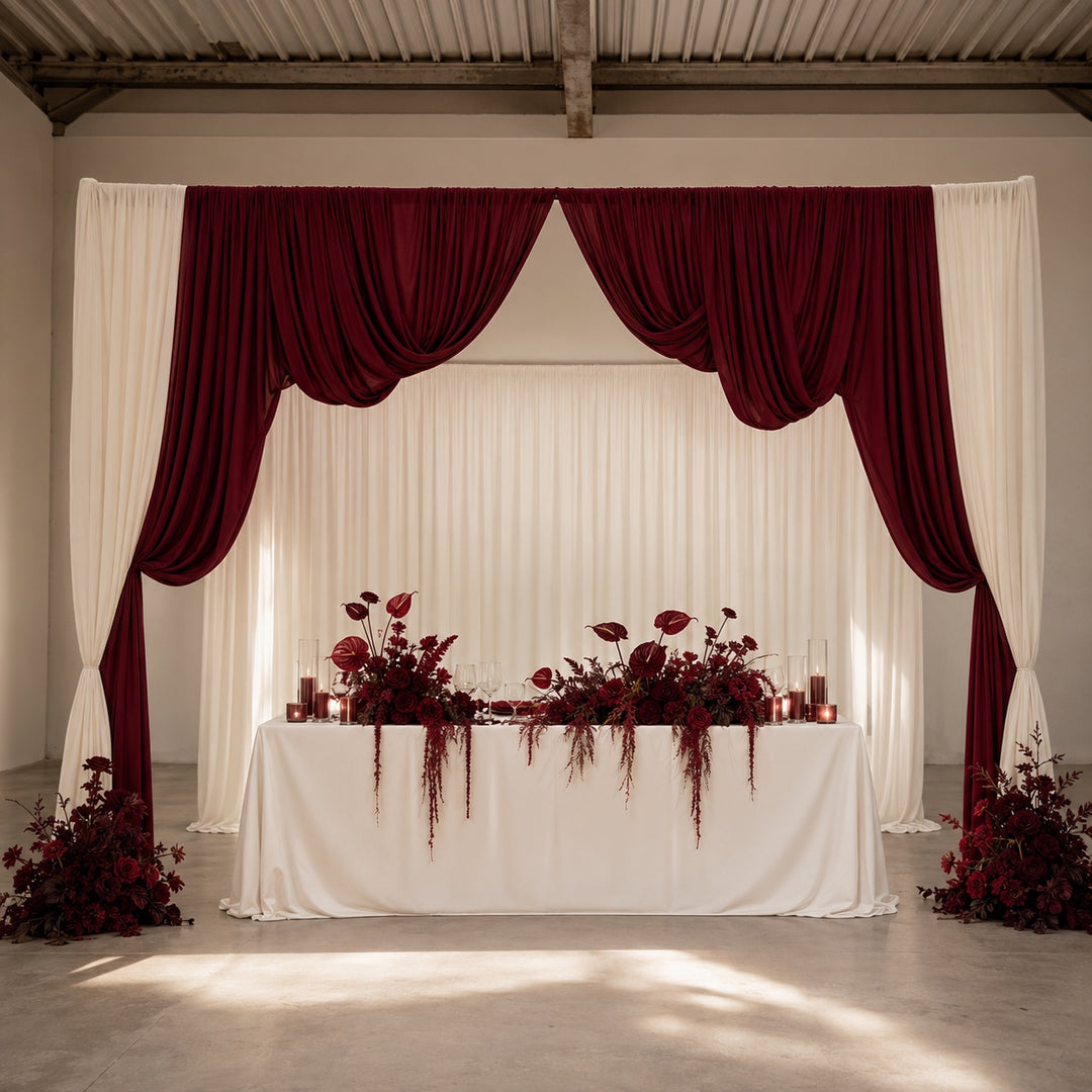A formal event backdrop featuring deep burgundy draped curtains and white drapes, with a white draped table decorated with matching burgundy floral arrangements in a bright, modern hall.