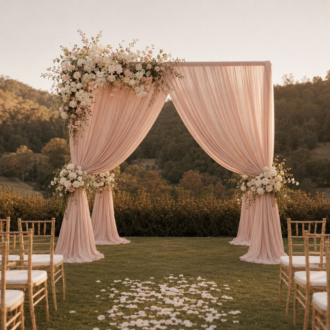 A peach-pink draped fabric wedding arch decorated with white and blush floral arrangements, set on a green lawn with scattered petals and wooden chairs, against a backdrop of hills at sunset.
