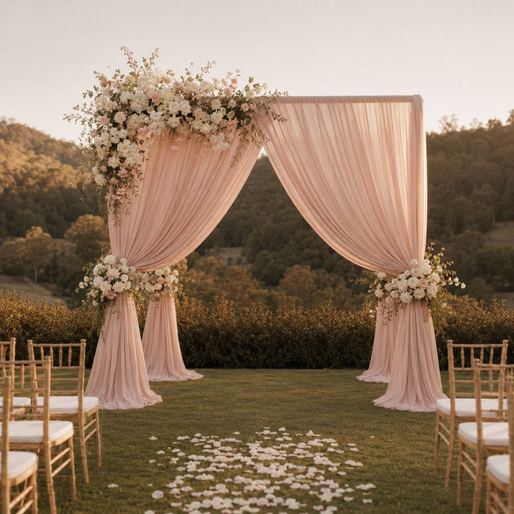 A peach-pink draped fabric wedding arch decorated with white and blush floral arrangements, set on a green lawn with scattered petals and wooden chairs, against a backdrop of hills at sunset.
