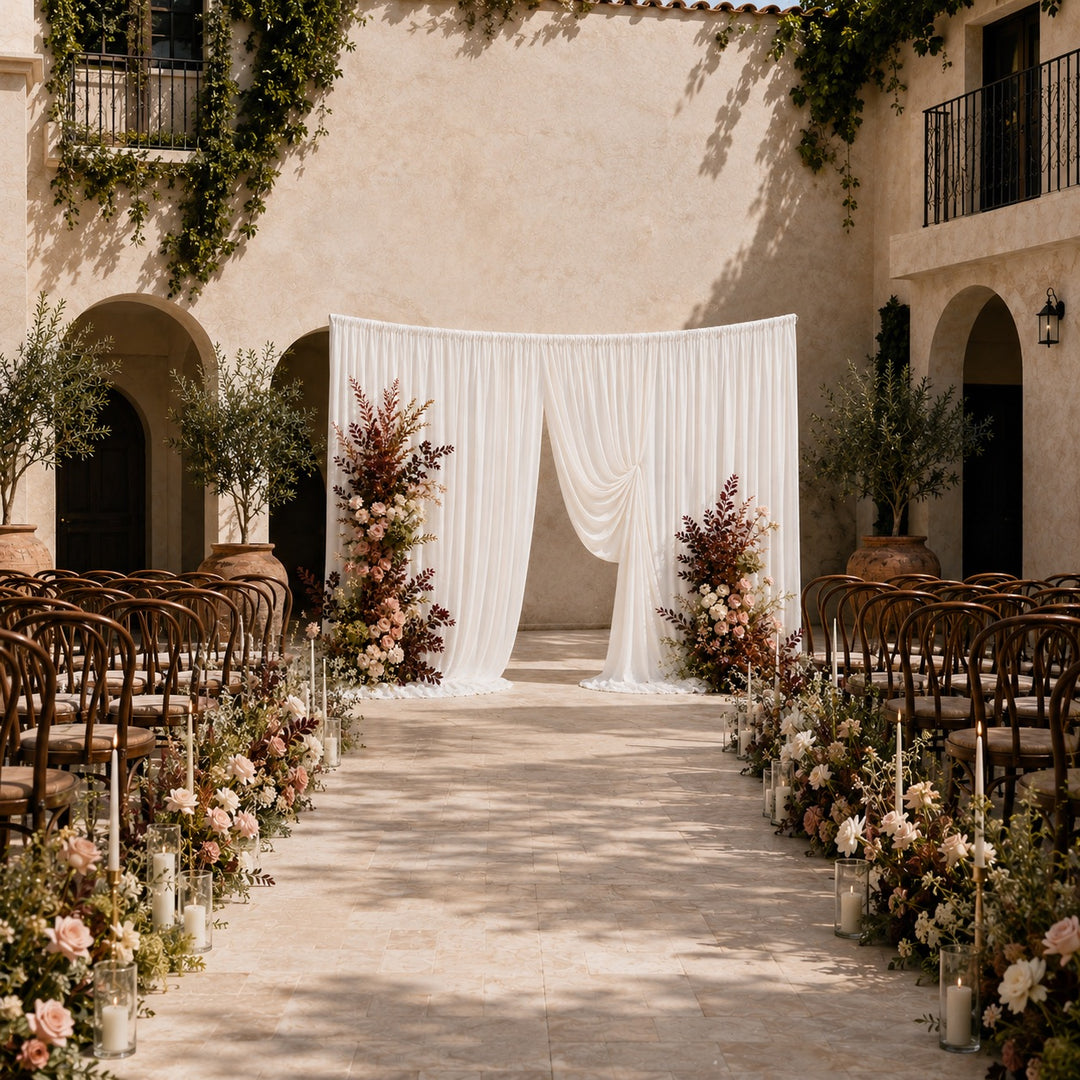 A wedding ceremony setup in a sunlit stone courtyard with rows of wooden chairs on both sides, floral arrangements along the aisle, and a white draped curved backdrop in the centre.