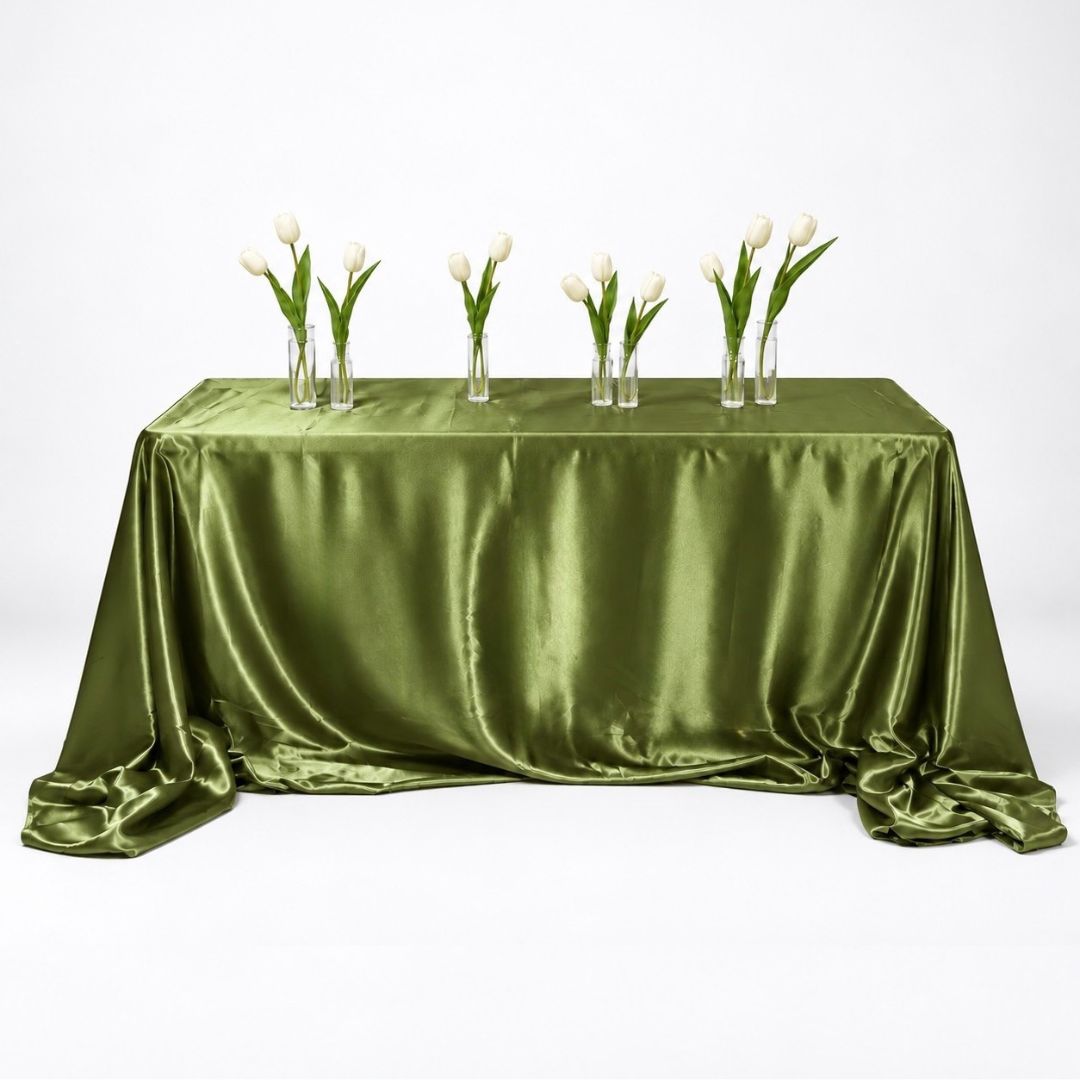 An olive green satin tablecloth draped over a rectangular table, styled with six clear glass vases containing white tulips, set against a seamless white studio background.