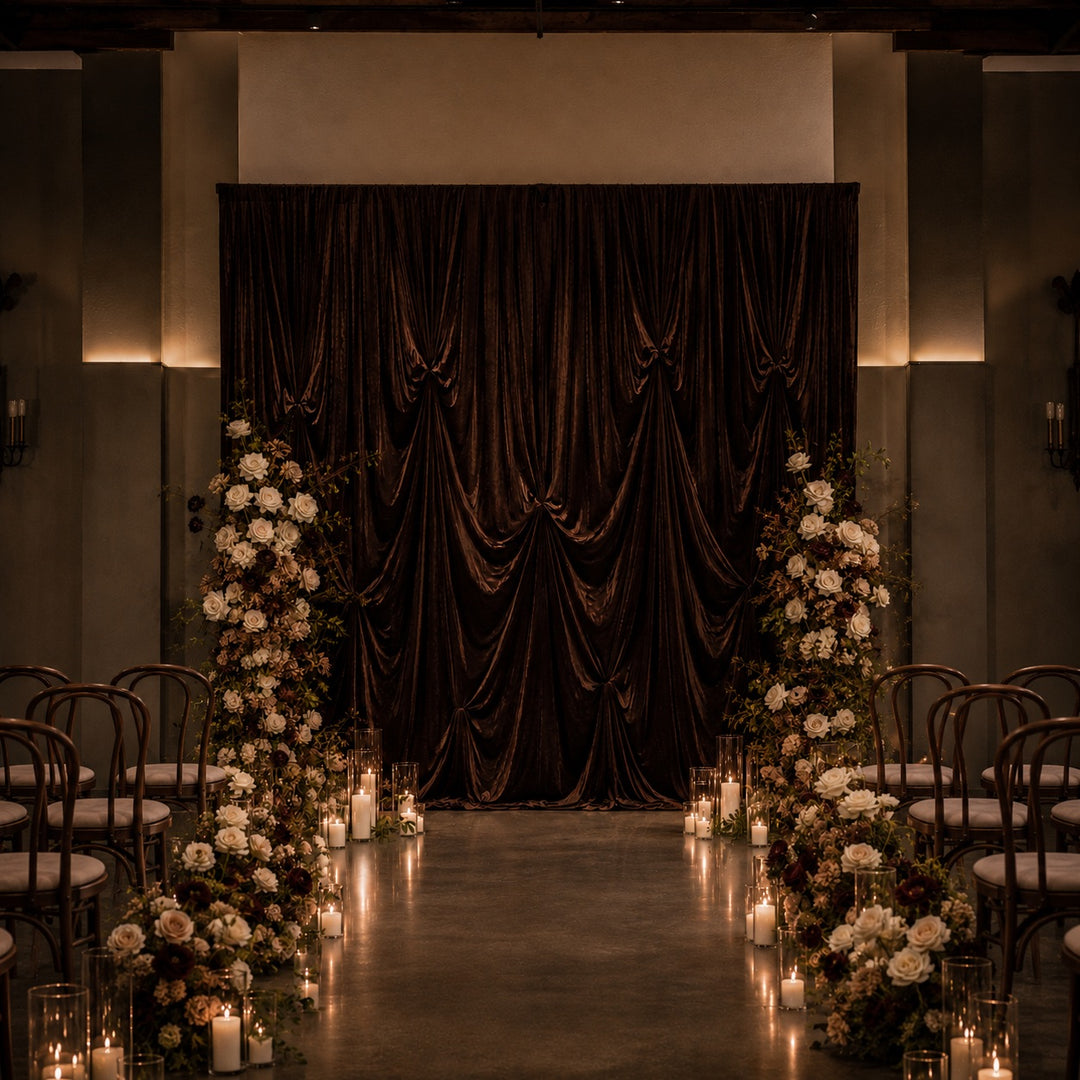 A dramatic event backdrop featuring a large draped dark brown velvet curtain, flanked by tall floral arrangements of white and brown flowers with lit candles on the floor, set against a dimly lit venue with rows of chairs.