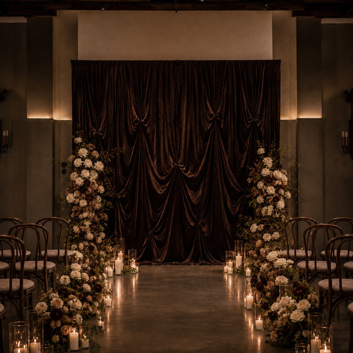 A dramatic event backdrop featuring a large draped dark brown velvet curtain, flanked by tall floral arrangements of white and brown flowers with lit candles on the floor, set against a dimly lit venue with rows of chairs.